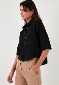 Black striped cropped shirt with a button front, short sleeves, and two chest pockets. Paired with beige pants, showcasing a neutral color palette.