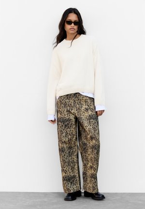 Tejfehér, bordázott pulóver, laza fazonban, széles, barna és fekete leopárdmintás nadrággal párosítva. Fekete napszemüveg és vastag fekete csizmák.