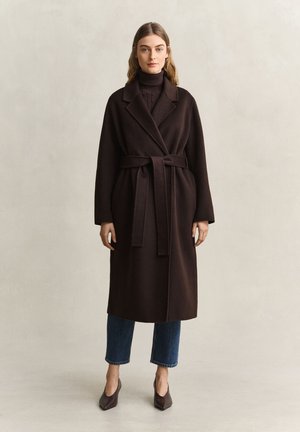 Femme portant un long manteau marron foncé ceinturé sur un pull à col roulé noir, un jean bleu court et des chaussures pointues marron, debout devant un fond uni.