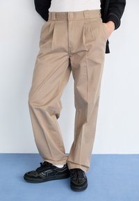 Pantaloni beige a pieghe realizzati in tessuto morbido, con vita alta e vestibilità rilassata, abbinati a sneakers nere con dettagli bianchi.