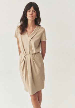 Beige short-sleeve dress with a v-neck, gathered waistband, and soft fabric. Features subtle pleats along the front for added texture.