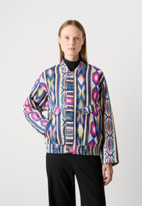 Vrouw in een kleurrijke, gestreepte jas met geometrische patronen en zwarte broek, staand met één hand in haar zak tegen een effen achtergrond.