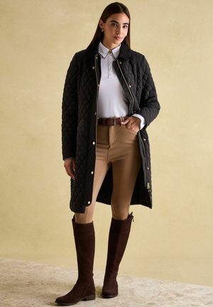 Quilted black long coat with snap buttons, white collared shirt, beige riding pants, and brown knee-high boots. Smooth texture, classic design.
