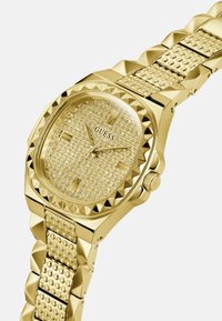 Reloj dorado con una esfera octagonal, adornada con cristales. Presenta una correa texturizada con acentos en forma de pirámide y un acabado pulido.