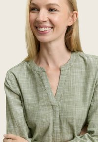 Blouse à manches longues à carreaux verts avec un décolleté en V. Le tissu présente un tissage léger et texturé. La mannequin a les cheveux longs et sourit.