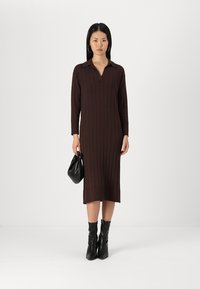 Brun, ribbet strikket kjole med lange ærmer og krave. Midtlængde med en tætsiddende silhuet. Sort håndtaske og sorte støvler med hæl som accessories.
