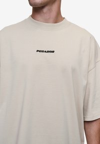 Béžové tričko s krátkym rukávom z bavlny, s čierne vyšitým logom "PEGADOR" na hrudi. Vo voľnom strihu s okrúhlym výstrihom.