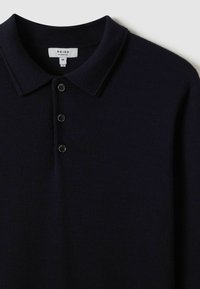 Polo din tricot de culoare bleumarin, cu o textură moale, trei butoane și un guler clasic pentru o ajustare perfectă. Eticheta brandului indică "REISS", mărimea M.