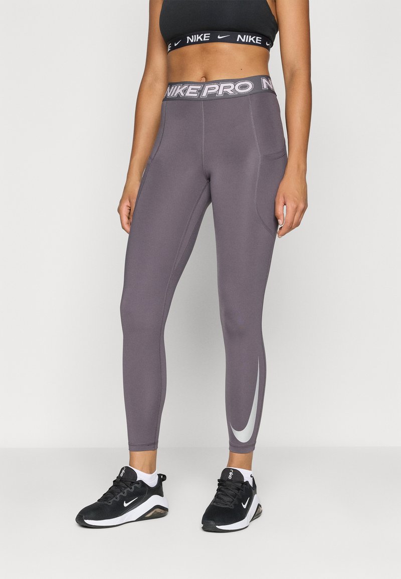 Nike Pro κολάν σε σκούρο γκρι χρώμα με σχέδιο ασημένιου swoosh στο πλάι. Ψηλή ζώνη με λογότυπο "NIKE PRO". Φορεμένα με μαύρα αθλητικά παπούτσια.