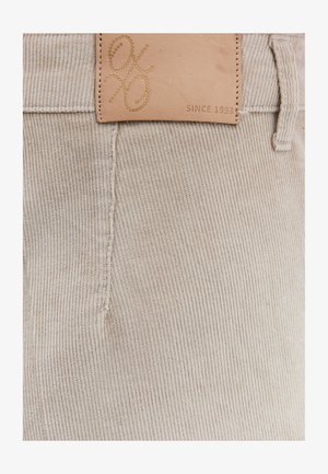 Beige kordfløjlstof med lodret ribstruktur, der har en tanfarvet læderlap med præget logo og teksten "SINCE 1993" nær linningen.