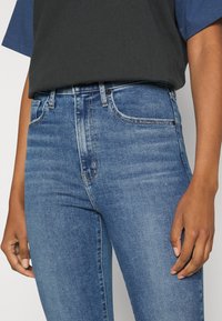 Vaqueros de mezclilla azul con silueta ajustada, cinco bolsillos y costuras visibles. Textura suave, cintura media y diseño clásico.