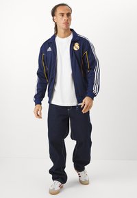 Veste zippée bleu marine avec accents jaunes et logo du Real Madrid ; associée à un jean en denim noir ample et des baskets blanches avec des rayures rouges.