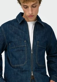 Jeans jakna temno modre barve, z zadrgo spredaj, dvema velikima žepoma na prsih in kontrastnim rumenim šivanjem na šivih in robovih.