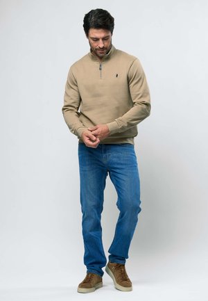 Mand justerer ærme iført beige sweatshirt med kvart-lynlås, blå jeans og brune sneakers mod en ensartet lys baggrund.