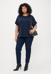 Tamnoplavi kratki rukav s opuštenim krojem, uparen s tamnim trapericama i crnim cipelama na petu, drži malu crnu torbicu preko ramena.