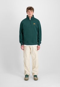 Felpa zip con colletto alto di colore verde scuro e piccolo logo sul petto, abbinata a pantaloni cargo beige chiaro e sneakers a fantasia.