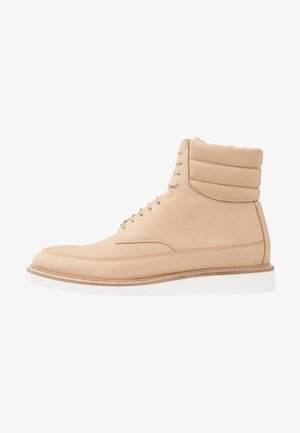 Bottes cheville en cuir beige clair avec col rembourré, fermeture à lacets à l'avant et semelle en caoutchouc blanche, présentant une texture lisse et un design minimaliste.