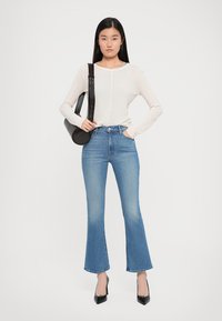 Lichtcrème langemouwen top met een ronde halslijn, gecombineerd met hoog getailleerde blauwe flared jeans. Een zwarte handtas en hakken maken de outfit compleet.