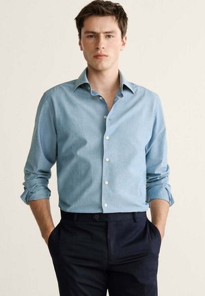 Joven de pie con las manos en los bolsillos, vistiendo una camisa azul clara con botones y mangas remangadas, y pantalones oscuros.