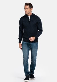 Man loopt gekleed in een donkerblauwe ritstrui van kabelbreiwerk, blauwe jeans en zwarte casual schoenen tegen een witte achtergrond.