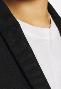 Černý blazer s límcem přes bílé tričko, které ukazuje texturovanou tkaninu s hladkým povrchem. Okraje límce a trička jsou jasně viditelné.