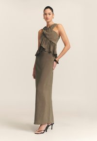 Olijfgroene jurk met asymmetrische rimpeldetails, getailleerde silhouet en een halternek. Draag met zwarte bandjeshakken.