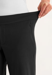 Zwarte broek met een zachte structuur, met een zijrits zak en een elastische tailleband, vastgehouden door een hand.