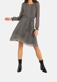 Vestido de patrón en blanco y negro con mangas largas, escote tipo llave y cintura ajustada con lazo. Combinado con botas de tobillo negras, tiene una silueta fluida.