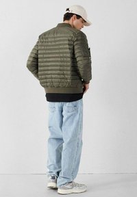 Giacca puffer verde oliva con design a righe orizzontali, orlo a coste e tasca con zip sulla manica, abbinata a jeans blu chiaro e sneakers.