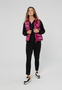 Gevoerde mouwloze vest in glanzend fuchsia met ritsluiting en logodetail, gecombineerd met een zwart top met rits en slanke zwarte broek, afgemaakt met sneakers.