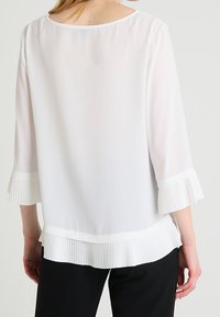 Valkoinen, kevyt paita, jossa on kolmannen neljänneksen pleidatut kellohihat ja pleidattu helma, sekä pyöreä kaula-aukko ja läpivalaistu kangas.