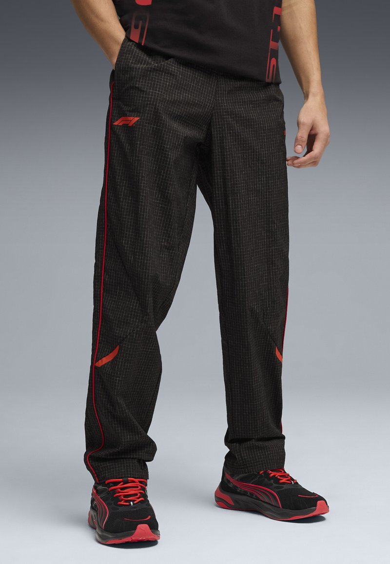 Homme portant un pantalon décontracté noir avec des bandes latérales rouges et des baskets noires avec des accents rouges, debout avec une main dans la poche.