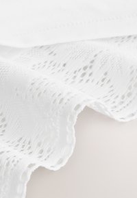 Tissu blanc avec un motif de dentelle complexe et un bord festonné, présenté en gros plan avec des plis doux et une texture visible.