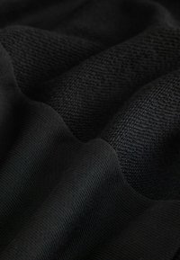 Svart tyg med varierade texturer; släta ribbade sektioner och en mer texturerad stickning som skapar ett lager på lager, vågigt utseende.