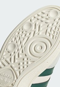 Podeszwa sneakersów Adidas z teksturowanym, wzorowanym designem z okręgami i geometrycznymi kształtami w kolorze kremowym, akcentowana ciemnozielonymi paskami.