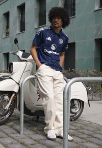 Modrý dres Adidas s bílými pruhy, krémové volné kalhoty a bílé tenisky. Vedle zaparkovaný bílý skútr, pozadí tvoří městská kamenná dlažba.