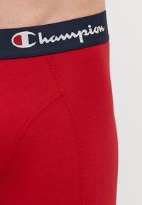 Crvene pamučne bokserice s mornarsko plavim elastičnim pojasom s bijelim "Champion" logotipom, teksturirana tkanina i kontrastni detalji šivanja.