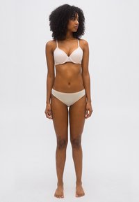 Γυναίκα με σγουρά μαλλιά, όρθια ξυπόλητη, φορώντας μπεζ σουτιέν και εσώρουχα, κοιτάζοντας προς τα αριστερά της μπροστά σε απλό ανοιχτόχρωμο φόντο.