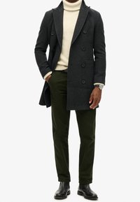 Grauer Wollmantel mit doppelter Knopfreihe über einem cremefarbenen Pullover, kombiniert mit olivgrünen Hosen und schwarzen Stiefeletten.