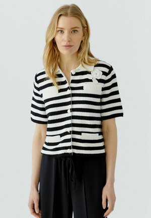 Vrouw met blond haar die een zwart-wit gestreept kort mouwen blouse met knopen, zakken en een kleine bloemdetail op de kraag draagt.