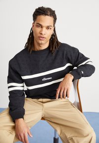 Felpa nera con strisce bianche, caratterizzata da un motivo texturizzato e un logo "ellesse" sul davanti. Modello seduto su una sedia.