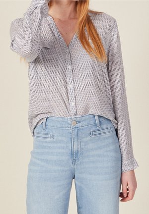 Lichte blouse met een geometrisch patroon in blauw en wit, met een V-hals, knoopsluiting aan de voorkant en lange mouwen. Gecombineerd met lichtblauwe jeans.