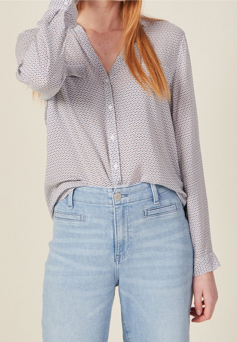 Lekka bluzka w geometryczny wzór w niebiesko-białych odcieniach, z dekoltem w kształcie litery V, zapinana na guziki z przodu i długimi rękawami. Połączona z jasnoniebieskimi dżinsami.