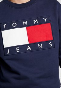 Sudadera azul marino con un gran logo que dice "TOMMY" en blanco y un diseño rectangular rojo y blanco debajo. Tejido suave y texturizado.