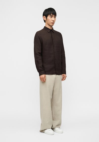 Καφέ λινό πουκάμισο με κουμπιά, μπεζ παντελόνι φαρδύ, λευκά αθλητικά παπούτσια· το μοντέλο στέκεται μπροστά σε απλό λευκό φόντο. Μινιμαλιστικός σχεδιασμός.