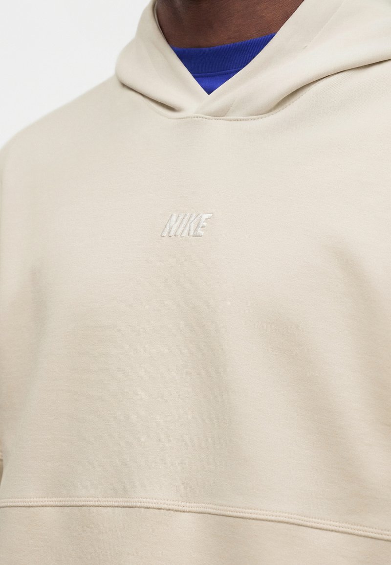 Nærbillede af beige Nike hættetrøje med præget logo på brystet, båret over en blå skjorte.