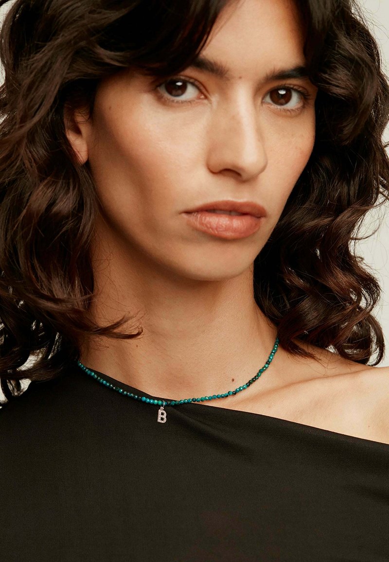 Collar de cuentas turquesas con un colgante de plata en forma de letra "B". Se muestra con una blusa oscura y cabello suave y ondulado enmarcando el rostro.