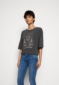 Camisa cinza de manga curta com uma impressão gráfica à frente com "Vibes Místicas", combinada com jeans azul de corte justo.