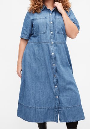 Femme portant une robe midi en jean bleu avec manches courtes, boutons-pression devant et deux poches poitrine, debout devant un fond uni.