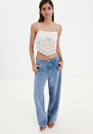 Femme debout, mains dans les poches, portant un crop top blanc brodé avec des bretelles fines et un jean bleu ample avec des sandales.